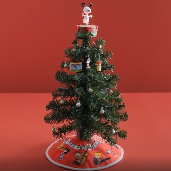 A miniature Christmas tree is topped with a Peanuts Snoopy tree topper that has light and plays music; there are miniature Peanuts Charlie Brown and Snoopy ornaments on the tree, and a matching tree skirt at its base.